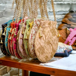 May include: A selection of embroidered drawstring bags in various colours, including black, teal, orange, pink, and peach. The bags feature floral embroidery, gold tassels, and are suspended from beaded cords. The bags are displayed on a wooden surface.