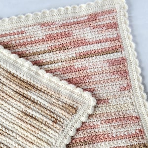 May include: A handmade crochet blanket in shades of pink, beige, and brown. The blanket has a striped pattern and a scalloped white border. The soft, textured blanket is suitable for a baby or as a decorative throw.