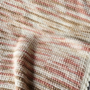 May include: A cream, light brown, and pink striped crochet blanket. The blanket features a textured stitch and a scalloped edge. This handmade blanket is ideal for adding warmth to a bed or sofa. The measurements are in centimetres.