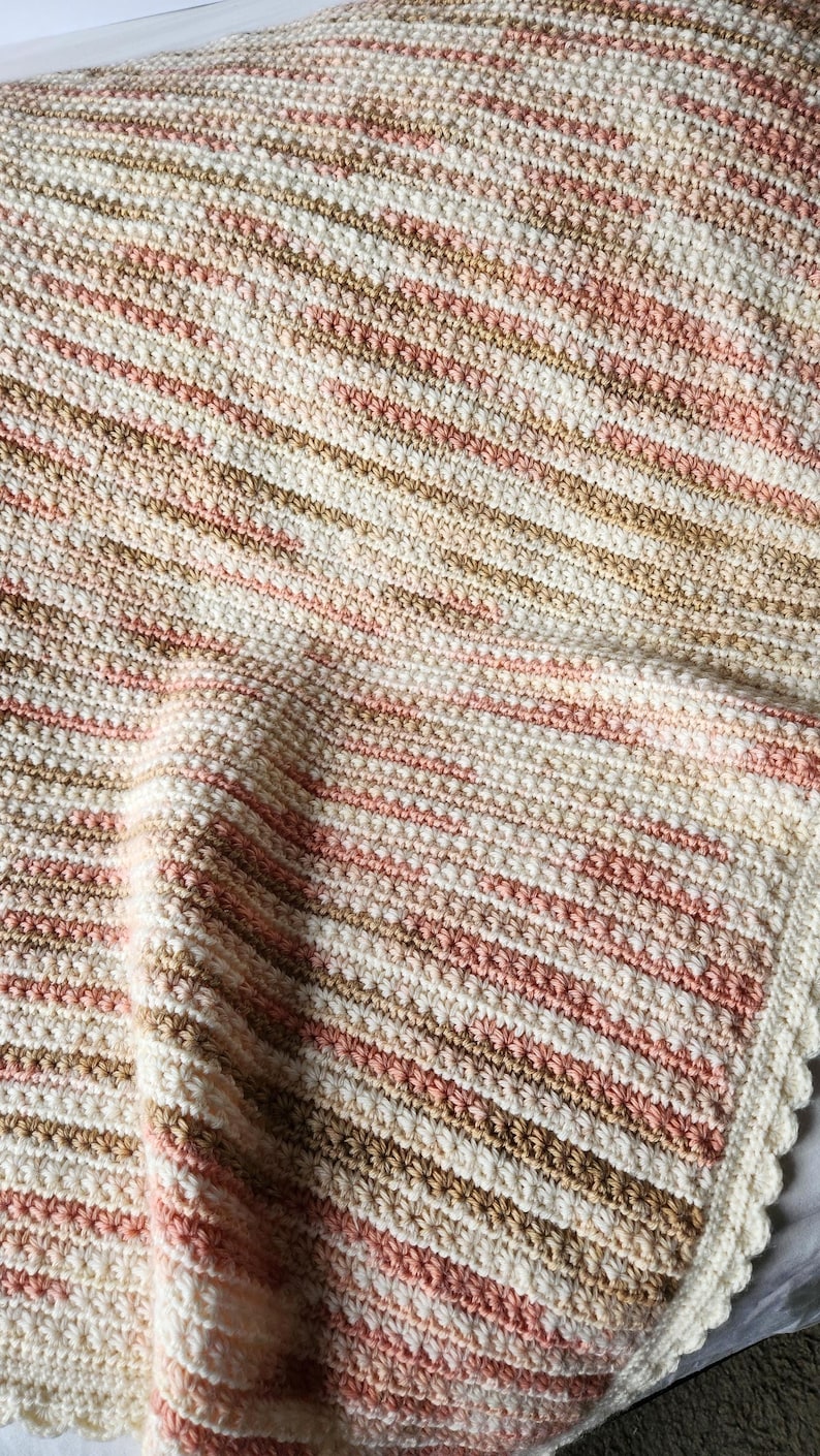 May include: A cream, light brown, and pink striped crochet blanket. The blanket features a textured stitch and a scalloped edge. This handmade blanket is ideal for adding warmth to a bed or sofa. The measurements are in centimetres.