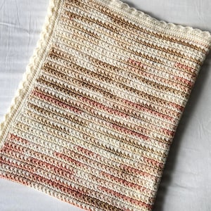 May include: A hand-crocheted blanket in cream, brown, and pink. The blanket has a textured stitch pattern with horizontal stripes and a scalloped cream border. This cosy blanket is ideal for adding warmth and style to any room.