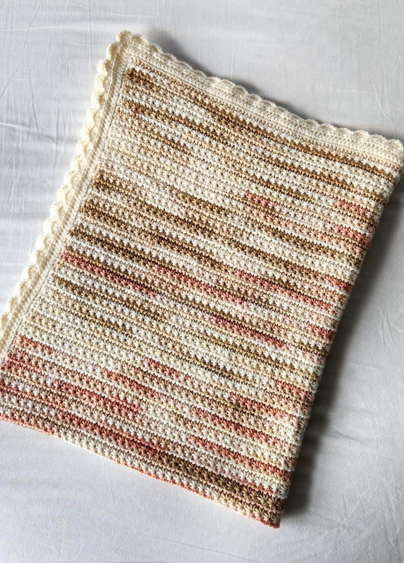May include: A hand-crocheted blanket in cream, brown, and pink. The blanket has a textured stitch pattern with horizontal stripes and a scalloped cream border. This cosy blanket is ideal for adding warmth and style to any room.