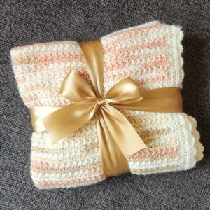 May include: A folded, handmade crochet blanket in cream, pink, and beige stripes, tied with a gold satin ribbon and bow. The blanket has a scalloped edge and is presented as a gift.