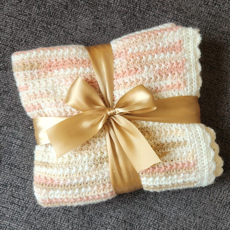 May include: A folded, handmade crochet blanket in cream, pink, and beige stripes, tied with a gold satin ribbon and bow. The blanket has a scalloped edge and is presented as a gift.