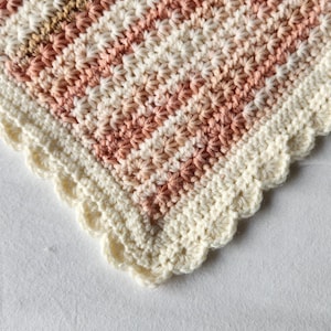 May include: Close-up of a handmade crochet blanket with a scalloped edge. The blanket features alternating stripes of cream, light brown, and shades of pink. The texture is detailed, showcasing the intricate stitch work. A cozy, decorative item.