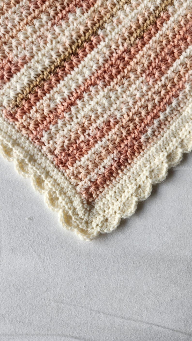 May include: Close-up of a handmade crochet blanket with a scalloped edge. The blanket features alternating stripes of cream, light brown, and shades of pink. The texture is detailed, showcasing the intricate stitch work. A cozy, decorative item.
