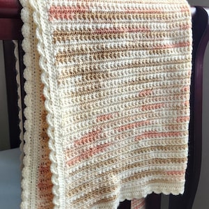 May include: A cream, peach, and tan crochet baby blanket with a scalloped edge, draped over a dark wood crib rail. The soft, textured blanket is a cosy nursery item, perfect for a newborn.