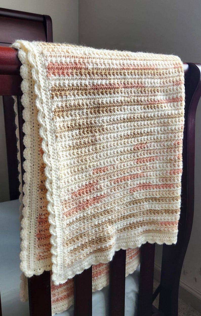 May include: A cream, peach, and tan crochet baby blanket with a scalloped edge, draped over a dark wood crib rail. The soft, textured blanket is a cosy nursery item, perfect for a newborn.