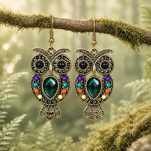 Könnte beinhalten: Ein Paar Eulen-Ohrringe in antikem Messing-Look mit filigraner Metallarbeit, schwarzen Perlenaugen und einem zentralen, smaragdgrünen facettierten Tropfenstein. Die Eulen sind mit kleinen, bunten Perlen in Violett, Blau, Orange und Gelb verziert.
