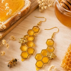 Può includere: Un paio di orecchini pendenti con una struttura a nido d'ape color oro riempita di resina gialla traslucida. Ogni orecchino è decorato con un piccolo ciondolo a forma di ape color oro dettagliato fissato al lato della cornice esagonale.