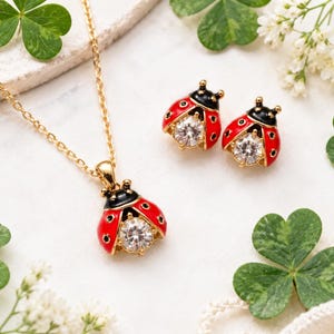 Pode incluir: Um conjunto de joias em tom dourado composto por um colar e um par de brincos. Cada peça apresenta um design de joaninha com as costas em esmalte vermelho brilhante, cabeça preta e uma pedra redonda, transparente e facetada no centro.
