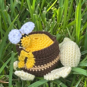 May include: Crocheted bee and honey pot set. The bee has white wings and a yellow and black body. The honey pot is brown with a yellow interior and a cream-colored stripe. The set is surrounded by green grass.