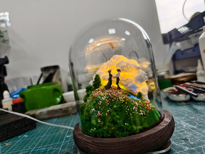 May include: A miniature diorama under a glass dome, featuring a couple standing on a green, mossy hill with colourful flowers. A glowing cloud-like structure provides a warm backdrop. The base is a dark wooden circle.