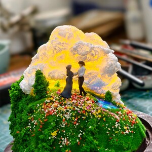 May include: A detailed miniature diorama depicting two figures on a grassy hill adorned with flowers. A luminous cloud-like structure provides a backdrop. The base is a circular wooden frame, creating a charming, artistic display.