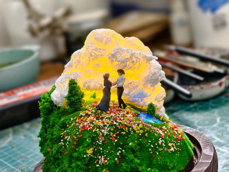 May include: A detailed miniature diorama depicting two figures on a grassy hill adorned with flowers. A luminous cloud-like structure provides a backdrop. The base is a circular wooden frame, creating a charming, artistic display.
