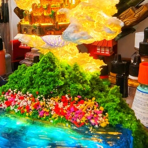 May include: A miniature fantasy landscape featuring a glowing castle atop a mountain. The mountain is covered in green foliage and colorful flowers, with a blue water feature in the foreground. The castle is illuminated with warm light.