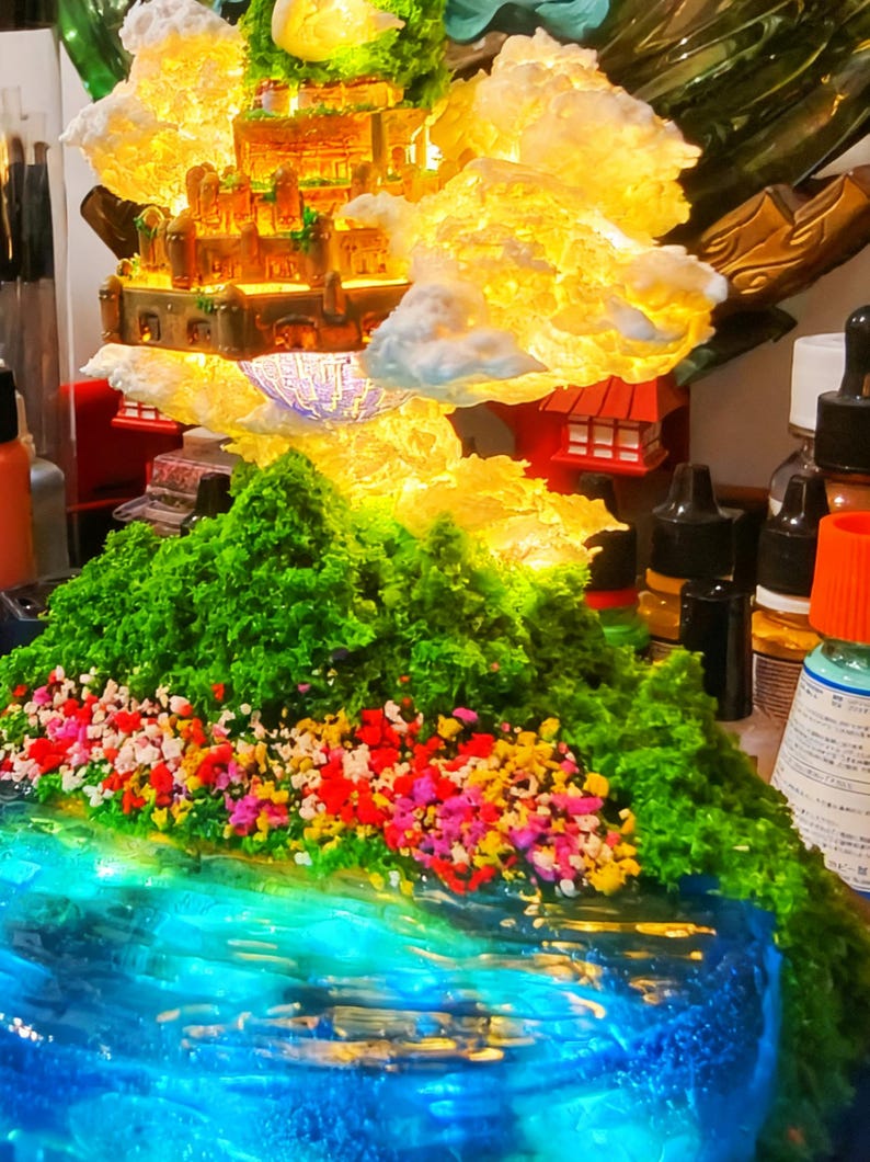 May include: A miniature fantasy landscape featuring a glowing castle atop a mountain. The mountain is covered in green foliage and colorful flowers, with a blue water feature in the foreground. The castle is illuminated with warm light.