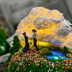 May include: A miniature diorama showing two figures holding hands on a grassy hill with colourful flowers. A glowing cloud formation illuminates the scene, with a small stream and trees. The figures are in a romantic pose.