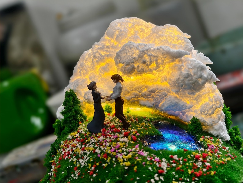 May include: A miniature diorama showing two figures holding hands on a grassy hill with colourful flowers. A glowing cloud formation illuminates the scene, with a small stream and trees. The figures are in a romantic pose.
