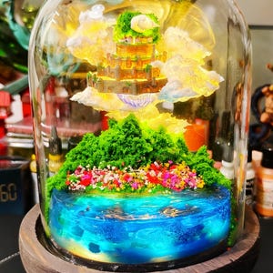 May include: A miniature fantasy landscape encased in a glass dome. The scene features a tiered castle, fluffy clouds, and a vibrant blue lake with colorful flowers and green foliage. The dome sits on a wooden base.
