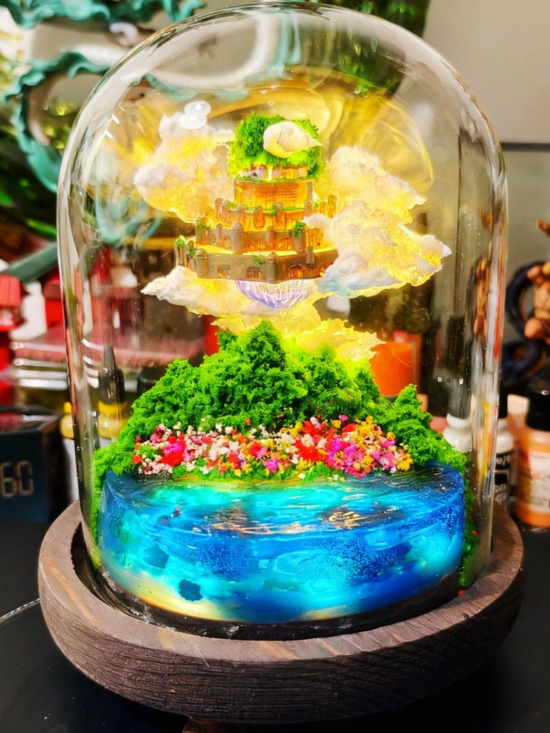 May include: A miniature fantasy landscape encased in a glass dome. The scene features a tiered castle, fluffy clouds, and a vibrant blue lake with colorful flowers and green foliage. The dome sits on a wooden base.
