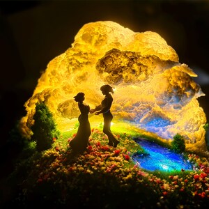 May include: A detailed diorama showing two figures silhouetted against a luminous, cloud-like backdrop. The scene includes a small pond and flowers, creating a whimsical, illuminated display. The figures are holding hands.