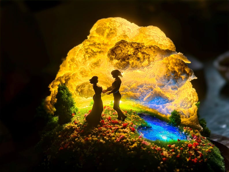 May include: A detailed diorama showing two figures silhouetted against a luminous, cloud-like backdrop. The scene includes a small pond and flowers, creating a whimsical, illuminated display. The figures are holding hands.