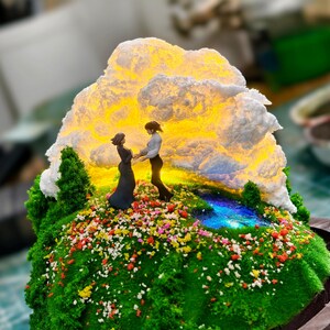 May include: A detailed miniature diorama depicting two figures on a grassy hill adorned with flowers, set against a glowing cloud. The base is a wooden frame, creating a scenic display.
