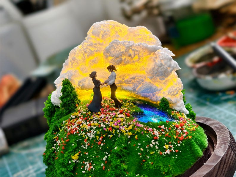 May include: A detailed miniature diorama depicting two figures on a grassy hill adorned with flowers, set against a glowing cloud. The base is a wooden frame, creating a scenic display.