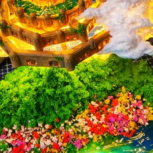 May include: A detailed miniature castle with illuminated windows and a surrounding landscape. The castle is brown with green accents, set against a backdrop of green foliage, colorful flowers, and a blue water feature. White fluffy clouds are also present.