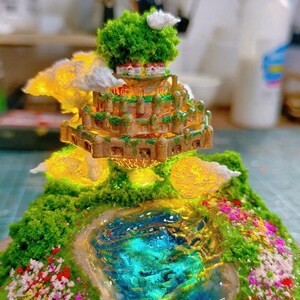May include: A miniature diorama lamp featuring a tiered structure with a green, mossy base, a blue pond, and colorful flowers. The structure has a castle-like appearance with a green tree on top. The text "Hanildmbole Diorama Lamp" is visible.