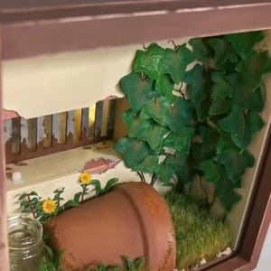 May include: A miniature diorama scene in a wooden frame. The scene includes a terracotta pot, green ivy, yellow flowers, and a small glass jar. The top of the frame has Japanese text.