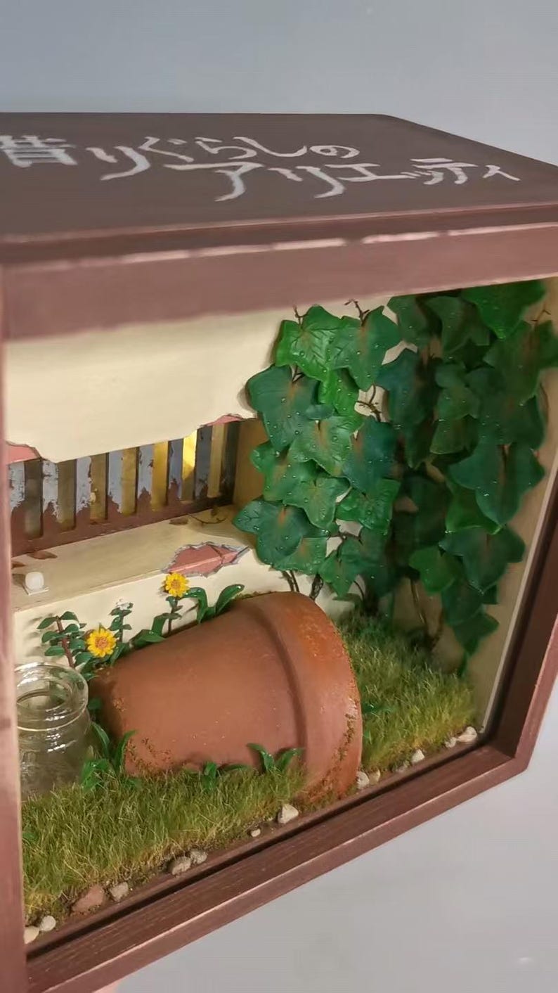 May include: A miniature diorama scene in a wooden frame. The scene includes a terracotta pot, green ivy, yellow flowers, and a small glass jar. The top of the frame has Japanese text.