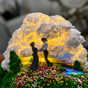 May include: A miniature diorama depicting two figures holding hands on a flower-covered hill. A glowing cloud-like structure illuminates the scene. The base is covered in green moss and colourful flowers, creating a romantic setting.