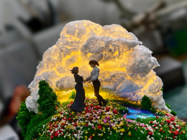 May include: A miniature diorama depicting two figures holding hands on a flower-covered hill. A glowing cloud-like structure illuminates the scene. The base is covered in green moss and colourful flowers, creating a romantic setting.