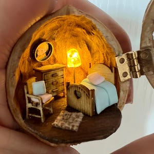 May include: A detailed miniature bedroom diorama housed within a walnut shell. The scene includes a bed with a blue blanket, a small chest of drawers, a chair, and a rug. A small framed picture is on the wall. A small lamp provides illumination.