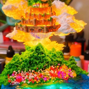 May include: A miniature fantasy landscape featuring a floating castle with detailed architecture, surrounded by fluffy clouds and illuminated by a warm glow. Below, a green, tree-covered island with colorful flowers sits above a blue, water-like base.