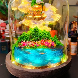 May include: A detailed miniature fantasy scene within a glass dome. It features a glowing castle, fluffy clouds, green hills, and a blue lake with colourful flowers. The base is a dark wooden platform.