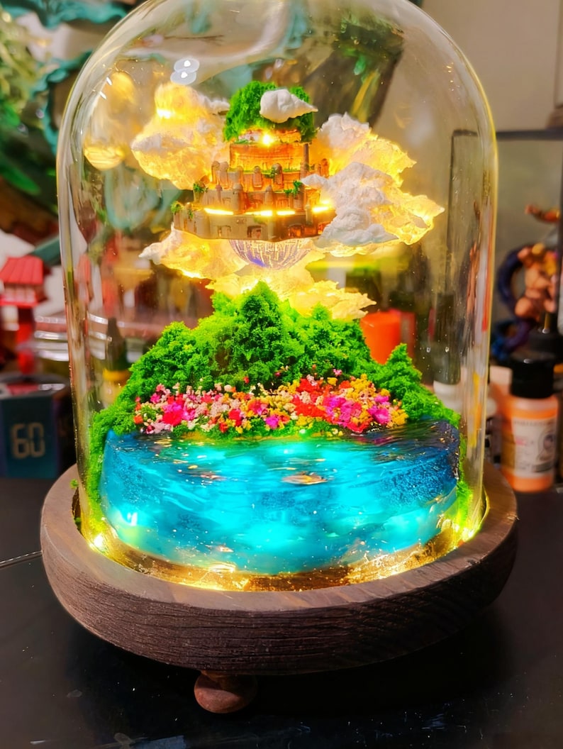 May include: A miniature fantasy landscape encased in a glass dome. The scene includes a glowing castle, fluffy clouds, green hills, and a blue lake with colorful flowers. The base is a dark wooden platform.