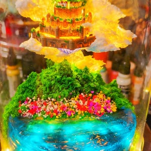 May include: A miniature fantasy landscape encased in glass. It features a tiered castle atop fluffy clouds, above a green, floral-covered island surrounded by a blue, glowing water feature. The scene is illuminated with warm, golden light.