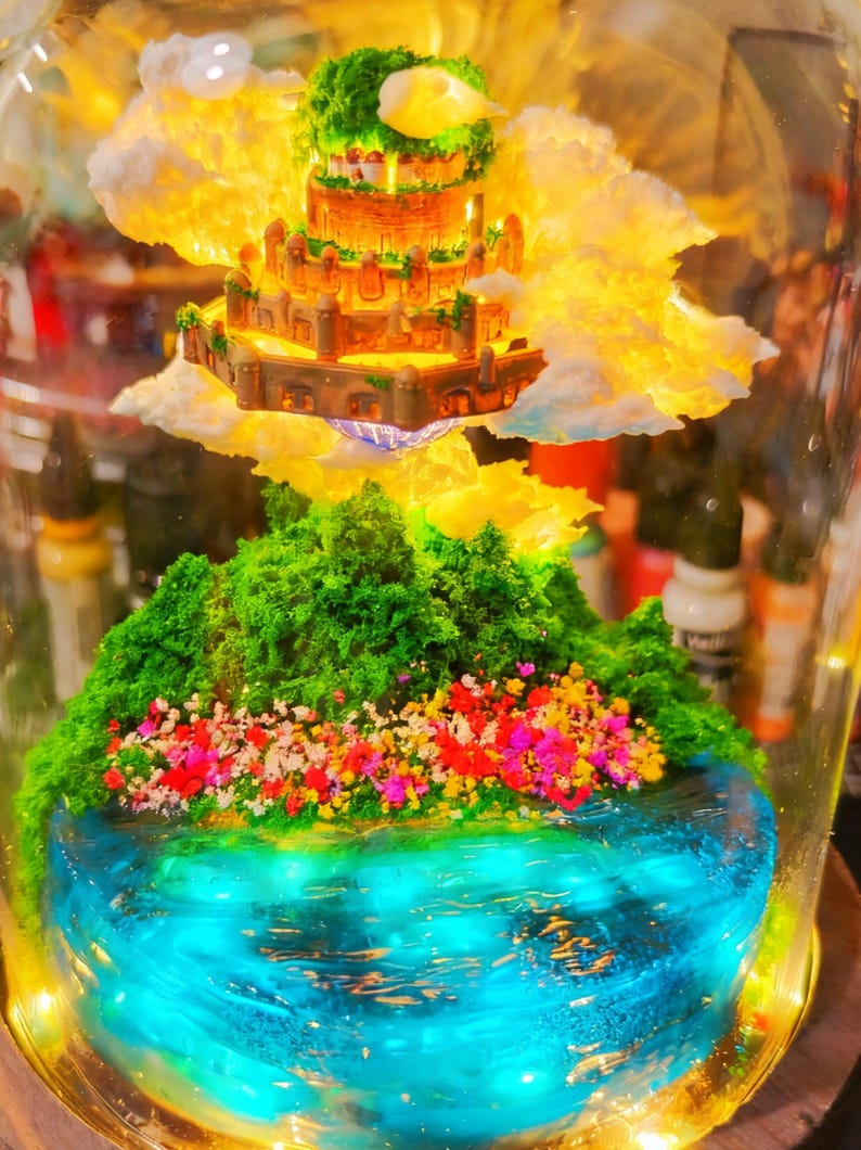 May include: A miniature fantasy landscape encased in glass. It features a tiered castle atop fluffy clouds, above a green, floral-covered island surrounded by a blue, glowing water feature. The scene is illuminated with warm, golden light.