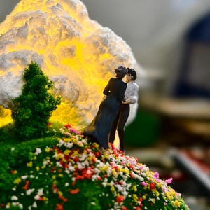 May include: A miniature diorama showing two figures embracing on a grassy hill with colourful flowers. A large, illuminated cloud-like structure glows in the background, with small green trees adding detail.