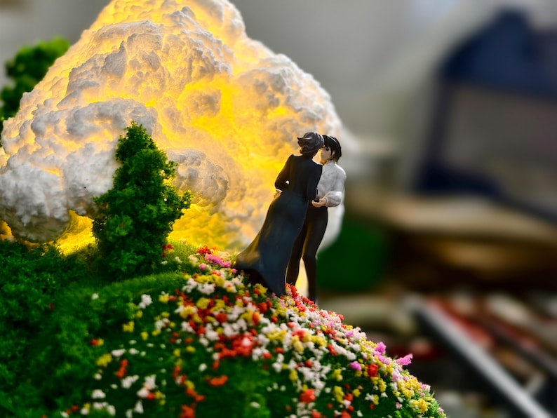 May include: A miniature diorama showing two figures embracing on a grassy hill with colourful flowers. A large, illuminated cloud-like structure glows in the background, with small green trees adding detail.