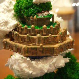 May include: A miniature fantasy castle with multiple tiers, detailed with small towers and windows. The castle is surrounded by green, textured foliage and fluffy white clouds, creating a whimsical, fairytale aesthetic.