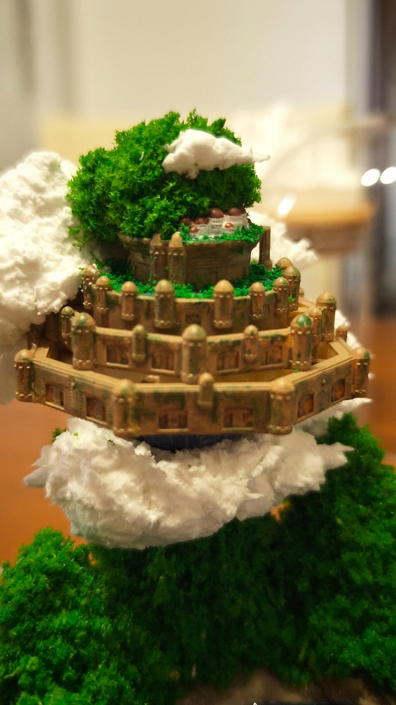 May include: A miniature fantasy castle with multiple tiers, detailed with small towers and windows. The castle is surrounded by green, textured foliage and fluffy white clouds, creating a whimsical, fairytale aesthetic.