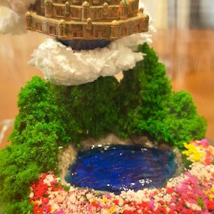 May include: Miniature fantasy landscape with a tiered, golden-brown structure, lush green foliage, and a vibrant blue pond. The scene is adorned with colorful flowers and fluffy white cloud accents, all set within a wooden base.