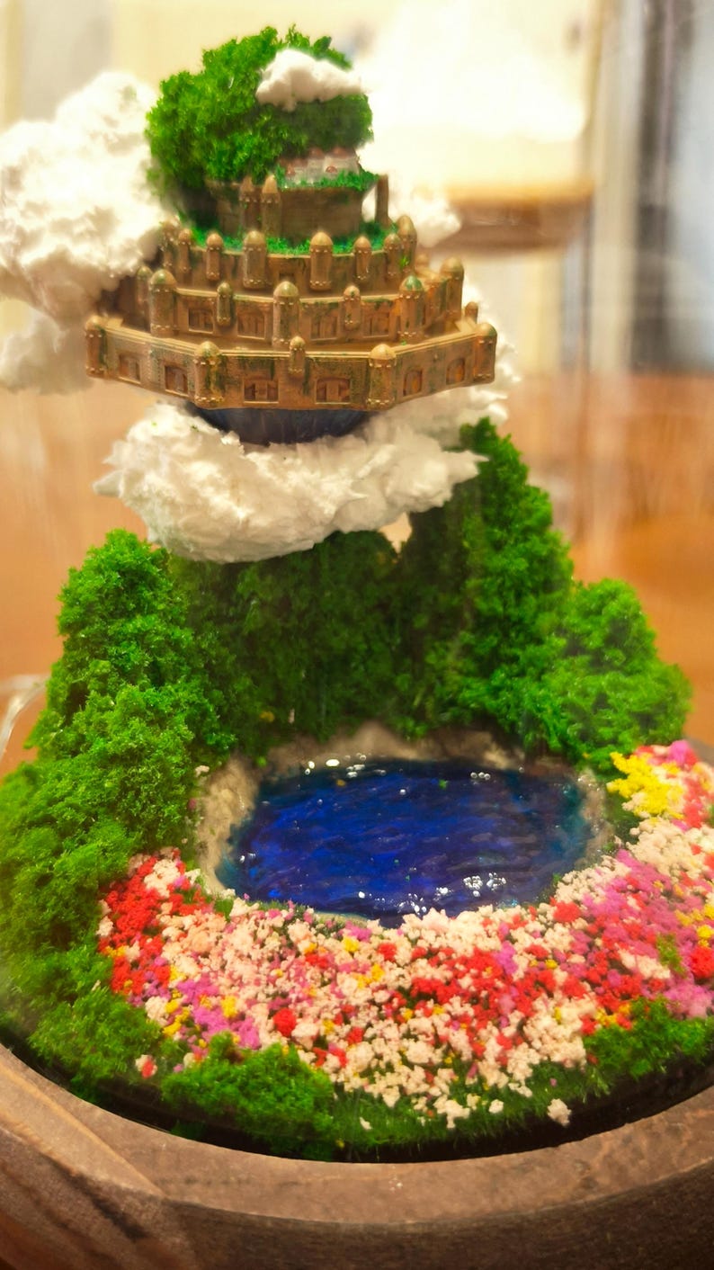 May include: Miniature fantasy landscape with a tiered, golden-brown structure, lush green foliage, and a vibrant blue pond. The scene is adorned with colorful flowers and fluffy white cloud accents, all set within a wooden base.