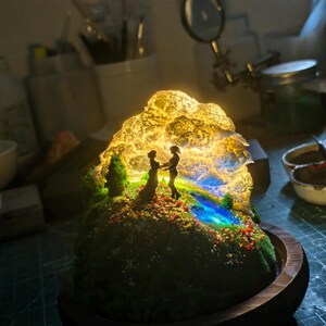 May include: A miniature illuminated diorama depicting a couple silhouetted against a glowing, cloud-like structure. The scene includes a miniature landscape with a stream, trees, and colourful flowers, set on a wooden base.