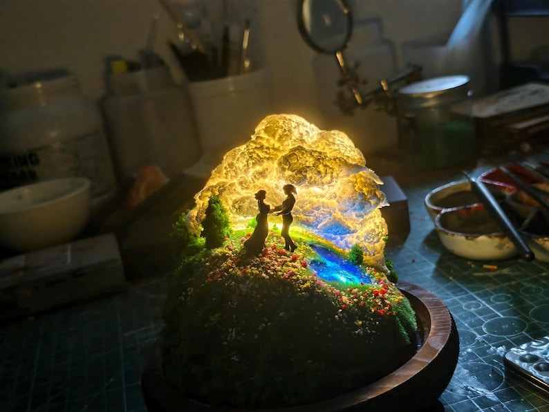May include: A miniature illuminated diorama depicting a couple silhouetted against a glowing, cloud-like structure. The scene includes a miniature landscape with a stream, trees, and colourful flowers, set on a wooden base.