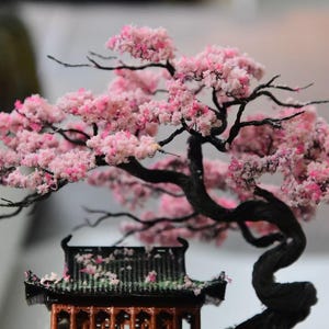 May include: Miniature bonsai tree with pink blossoms and a small wooden pagoda. The tree has a dark, twisted trunk and branches, with delicate pink flowers. The pagoda has a black roof and intricate details.